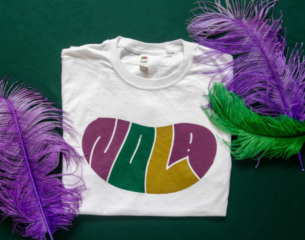 White T-shirt with "NOLA" in purple, green, and gold letters, set on a dark green surface. Flanked by purple and green feathers, evoking a festive vibe.