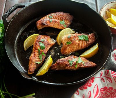 Pan-seared salmon fillets in a skillet, garnished with parsley and lemon wedges. Warm lighting and a red patterned cloth convey a cozy, appetizing atmosphere.