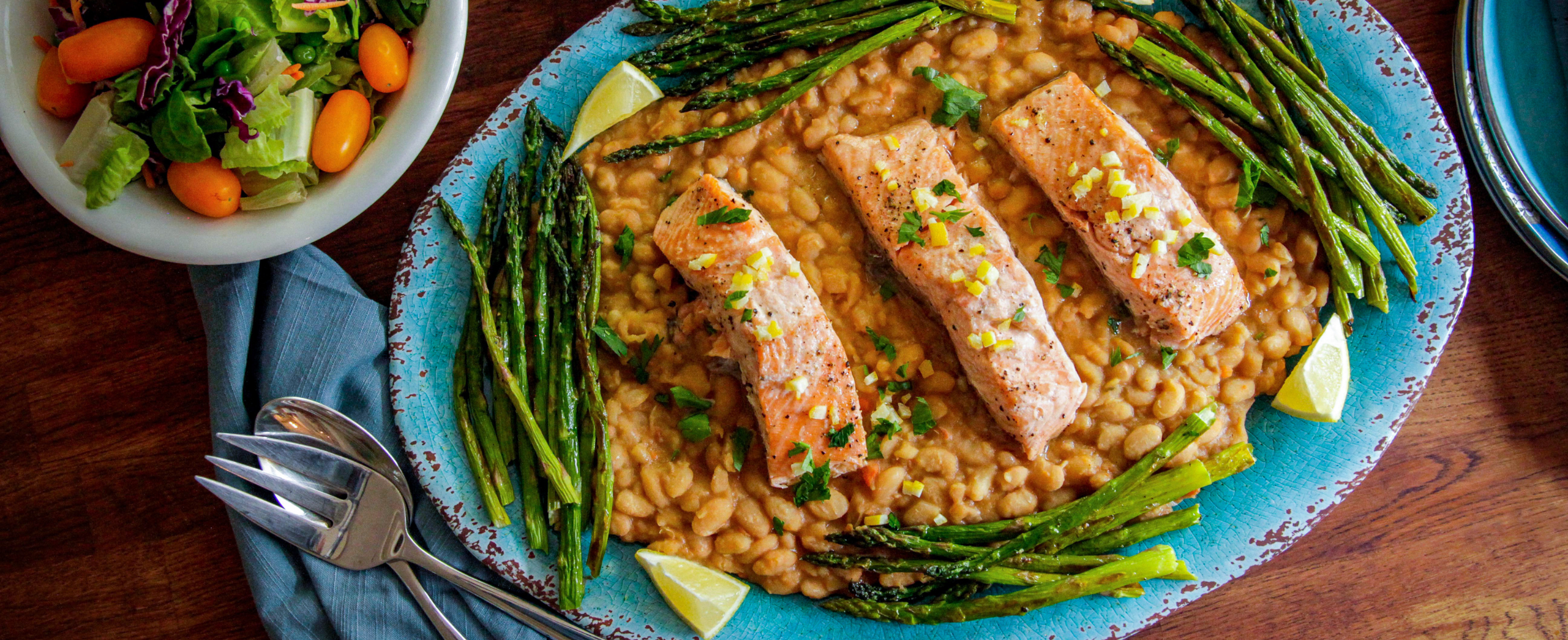 Three pieces of cooked salmon on a bed of beans, garnished with lemon wedges and grilled asparagus, served on a blue platter with a side salad and utensils nearby.
