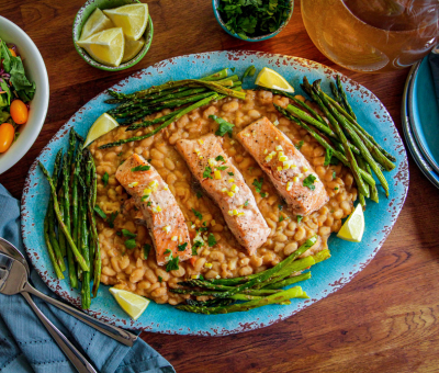 Three pieces of cooked salmon on a bed of beans, garnished with lemon wedges and grilled asparagus, served on a blue platter with a side salad and utensils nearby.