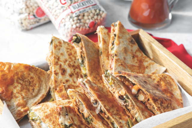 Golden quesadillas filled with beans and vegetables in a wooden tray, a pitcher of sauce, and bean bags in the background. Plates stacked on the side.