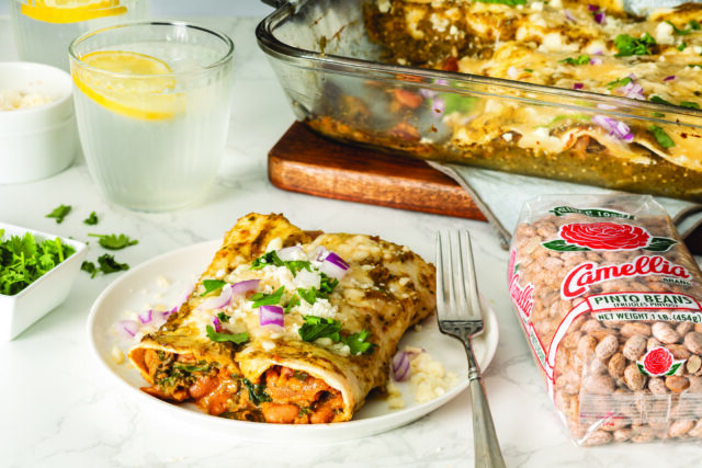 Two enchiladas on a white plate, topped with cheese, onions, and cilantro. A glass of lemon water and a bag of pinto beans are nearby, with a baking dish in the background.