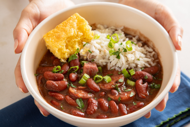Hands holding a white bowl of red beans and rice with sausage, topped with white rice, chopped green onions, and a square of cornbread.