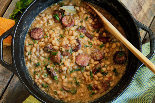 Cast iron skillet of white beans with sliced andouille sausage and fresh parsley, served with a wooden spoon on a rustic wooden table.