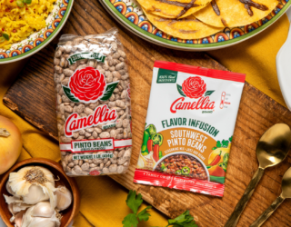 Camellia Pinto Beans and Southwest Pinto Beans Flavor Infusion on a wooden board with garlic, onion, and herbs, alongside cooked rice, tortillas, and a pot of beans.