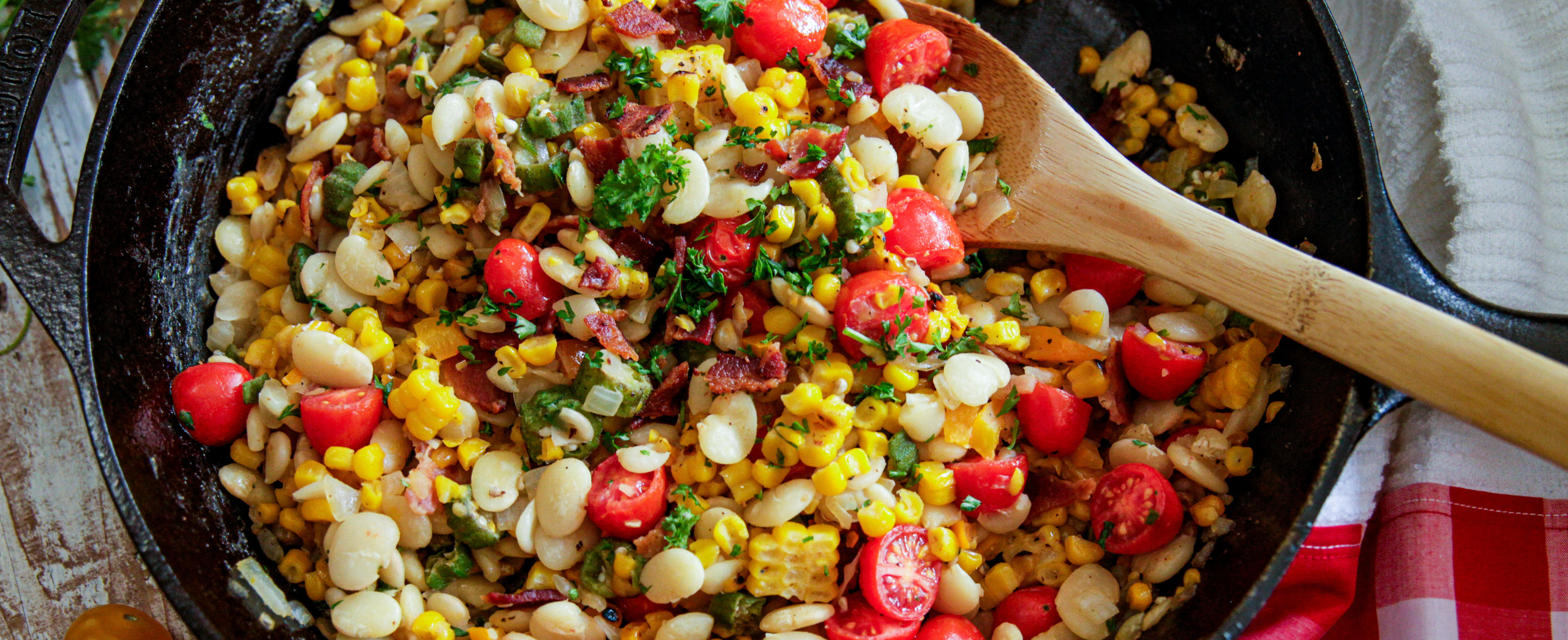 A colorful succotash in a skillet with corn, lima beans, cherry tomatoes, bacon, and parsley, stirred with a wooden spoon. A cheerful, fresh summer dish.