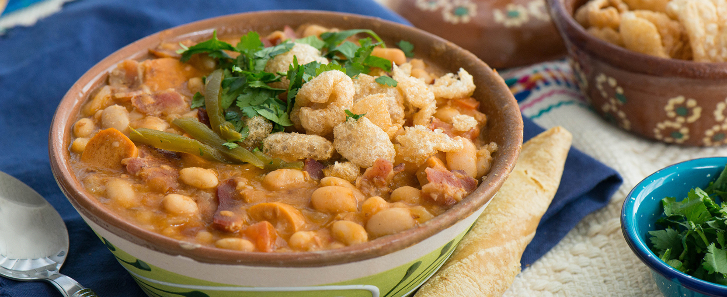 A bowl of hearty bean stew garnished with cilantro and crispy pork rinds, accompanied by bread on a blue cloth. Warm and comforting ambiance.