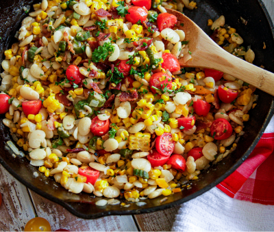 A colorful succotash in a skillet with corn, lima beans, cherry tomatoes, bacon, and parsley, stirred with a wooden spoon. A cheerful, fresh summer dish.
