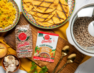 Camellia Pinto Beans and Southwest Pinto Beans Flavor Infusion on a wooden board with garlic, onion, and herbs, alongside cooked rice, tortillas, and a pot of beans.