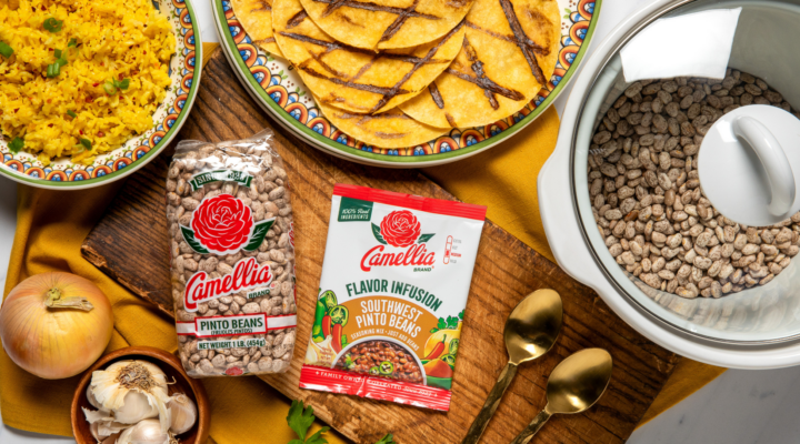 Camellia Pinto Beans and Southwest Pinto Beans Flavor Infusion on a wooden board with garlic, onion, and herbs, alongside cooked rice, tortillas, and a pot of beans.