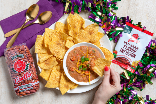 Hand dipping tortilla chip into bowl of red bean dip topped with cheese and green onions, surrounded by Camellia Red Kidney Beans bag, Red Bean Flavor Infusion packet, and Mardi Gras beads.