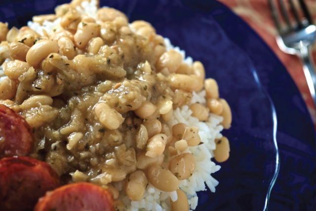White beans and rice over white rice on a blue plate with sausage, served alongside hot sauce.
