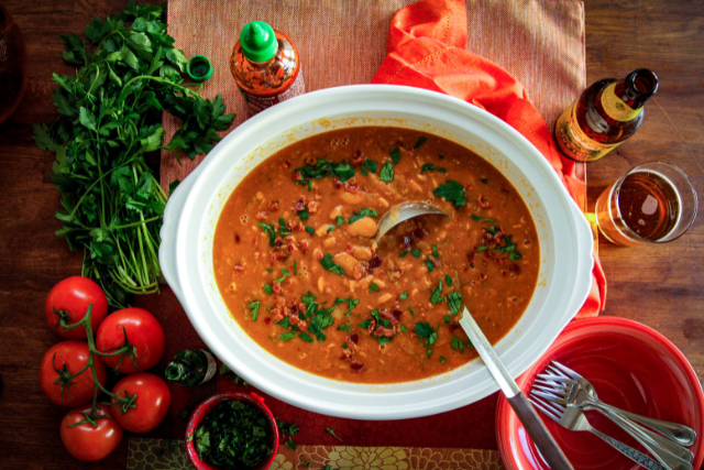 Bowl of lima beans garnished with fresh parsley, surrounded by fresh parsley bunch, tomatoes, hot sauce, and beer on a wooden table.