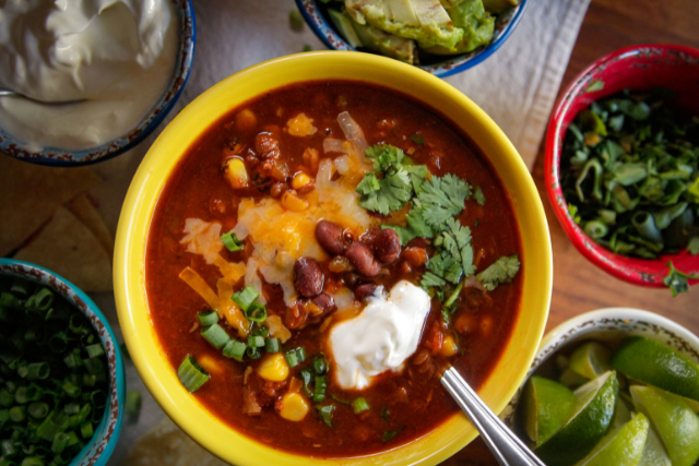 Taco soup in a yellow bowl topped with shredded cheese, sour cream, and cilantro, surrounded by bowls of toppings including avocado, lettuce, and lime wedges.
