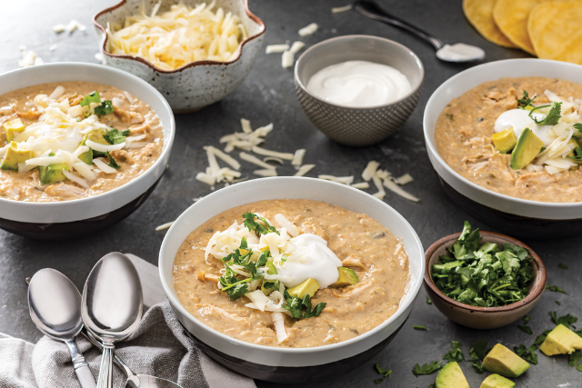 Three bowls of white chicken chili topped with sour cream, shredded cheese, cilantro, and avocado, served with crackers and garnish bowls.