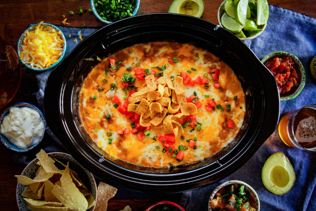 Slow cooker chicken tortilla soup topped with crushed tortilla chips, diced red peppers, and cilantro, surrounded by bowls of toppings including cheese, sour cream, avocado, and lime wedges.