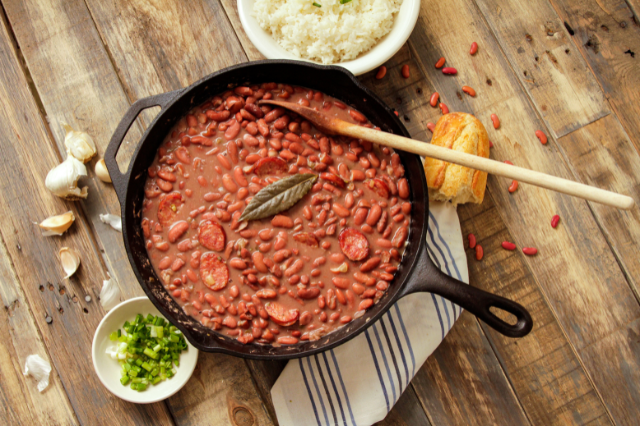 Cast iron skillet of red beans with sausage and bay leaves on a wooden table, surrounded by cooked rice, chopped green onions, garlic, and cornbread.