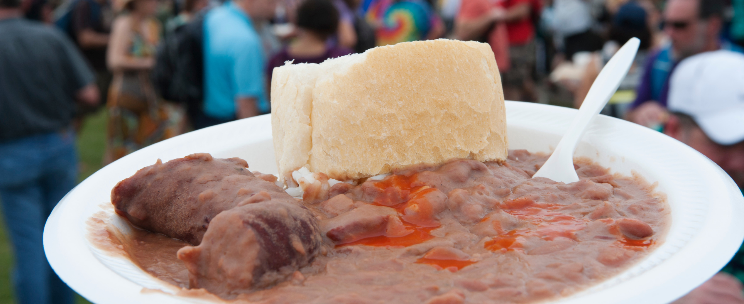 A white plate with sausage, beans in sauce, and a bread roll. In the background, a lively outdoor festival crowd is slightly blurred.