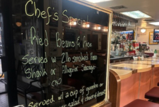 Chalkboard with "Chef's Special" menu featuring red beans and rice, at a cozy restaurant bar with warm lighting and bottles on shelves.
