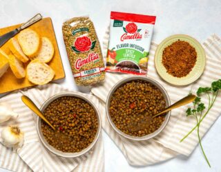 Overhead photo of bowls of cooked, Spicy Lentils, surrounded by bread, fresh garlic, bowl of spices, an unopened 1 pound bag of Lentils, and unopened Spicy Lentils Flavor Infusion package.