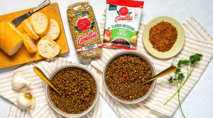 Overhead photo of bowls of cooked, Spicy Lentils, surrounded by bread, fresh garlic, bowl of spices, an unopened 1 pound bag of Lentils, and unopened Spicy Lentils Flavor Infusion package.