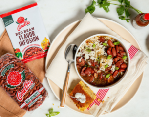 A bowl of red beans and rice garnished with green onions sits on a plate with cornbread. A package of red beans and seasoning are beside it, conveying warmth and home-cooked comfort.